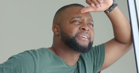 Leaning african man shading eyes and gazing out window, contemplative natural light