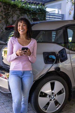 Woman Charging Electric Car While Using Smartphone Outdoors