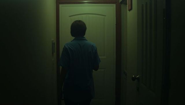Man standing facing paneled door in dim green hallway cinematic moody back view
