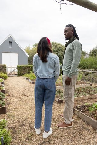 Diverse couple talking on gravel path through raised garden beds near gray outbuilding
