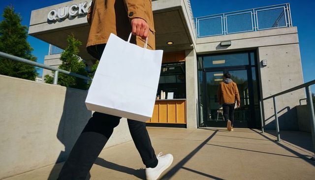 Carrying white shopping bag walking toward modern coffee shop entrance on urban sidewalk
