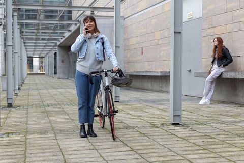 Diverse women standing in urban courtyard with bicycle while talking on smartphone