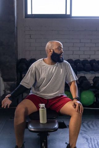 Bearded man resting on bench holding water bottle after workout in gym with dumbbells