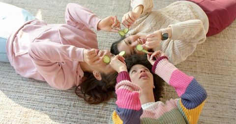 Diverse female friends lying on rug holding cucumber slices, wearing cozy sweaters during self-care