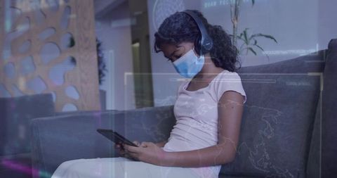 Young girl wearing face mask and headphones sitting on sofa using tablet in waiting lounge