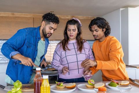 Indian friends cooking burgers in modern kitchen, flipping patties and assembling toppings