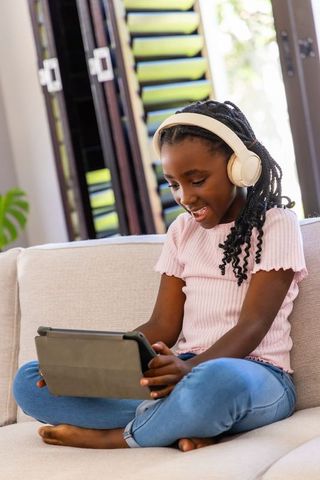 Young african american girl using tablet and wearing headphones on sofa learning at home