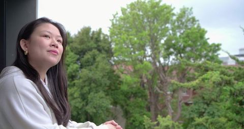 Asian woman leaning on balcony railing gazing at green trees, serene urban contemplation