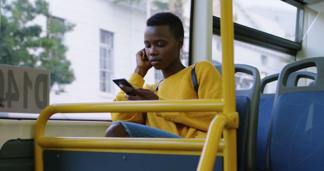 Young woman with short hair wearing a yellow sweater focused on using her smartphone during a bus ride in an urban area. Great for illustrating themes of urban lifestyle, public transportation, modern technology, and day-to-day commuting. Perfect for articles or advertisements related to public transit, tech usage, solo travel, or contemporary lifestyle.