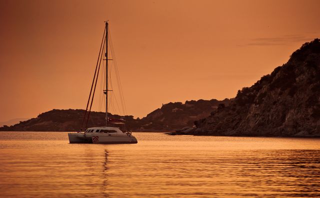 Sailboat anchored on calm sea during sunset with vivid orange sky and reflection on water. Ideal for travel and tourism, nature blogs, or background images promoting relaxation and vacations.