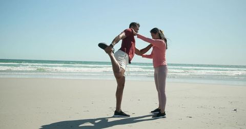Diverse couple stretching on sunny beach before run fitness session by ocean horizon