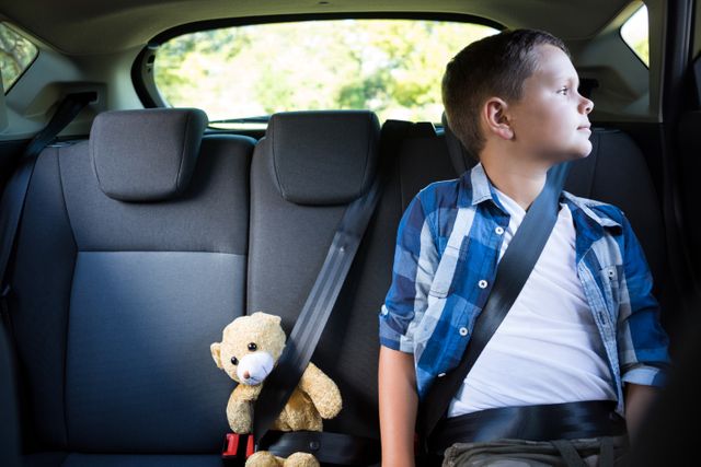 Boy sitting in the back seat of a car with a teddy bear beside him, both secured with seatbelts. He is looking out the window, suggesting a road trip or family travel. Ideal for use in articles or advertisements about child safety, family travel, or childhood memories.