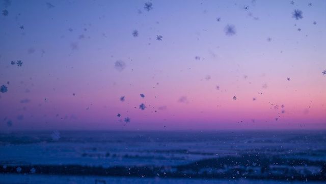 Frosted window with delicate snowflakes falling against pastel sunset over ocean horizon