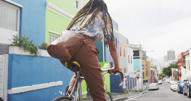 Rear view of a biracial man with long dreadlocks out and about in the city on a sunny day, wearing headphones, riding his bicycle in the street in slow motion.