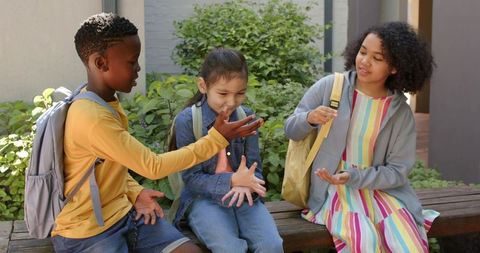 Multicultural students laughing and sharing backpacks while sitting on campus bench