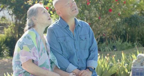 Senior asian couple sitting on tiled wall in sunny garden looking up with calm companionship
