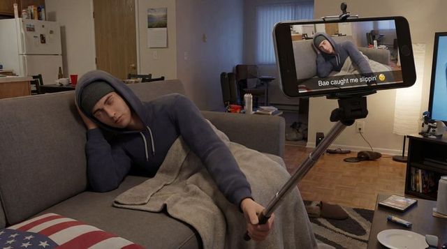 Young man sleeping on couch holding selfie stick recording prank for social media