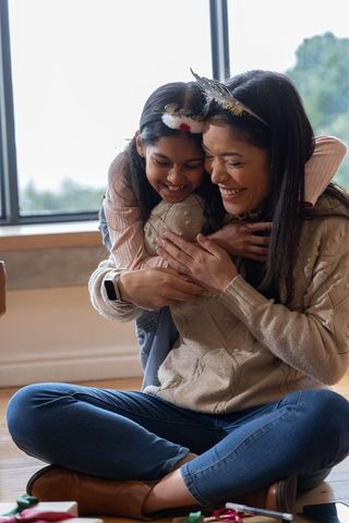 Mother and daughter sharing cozy hug by window, smiling and laughing over gifts
