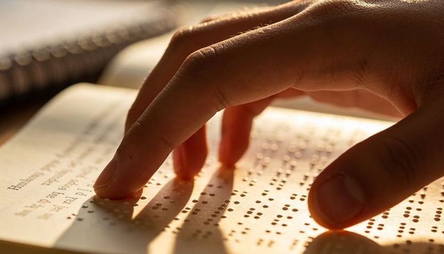 Right hand reading braille page in warm light showing tactile dots and fingertip motion