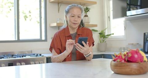 Senior asian woman using smartphone at marble kitchen island with fruit bowl and plants
