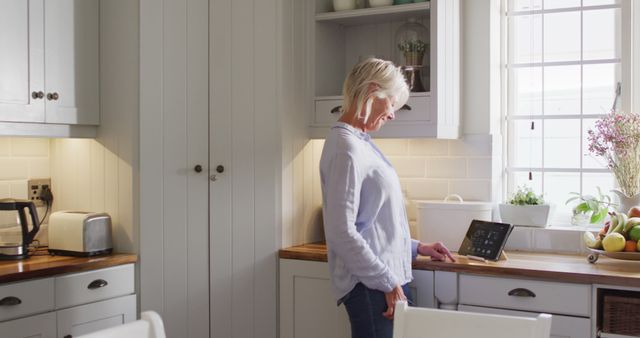 Happy senior caucasian woman standing in kitchen, using smartphone and tablet. Spending quality time at home alone.