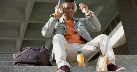 Young man adjusting headphones on urban steps with backpack and juice bottle denim jacket sneakers