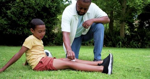 African american father kneeling cleaning son's scraped knee after backyard soccer play