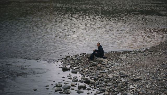 Solitary man sitting on rocky shoreline watching calm lake under overcast sky