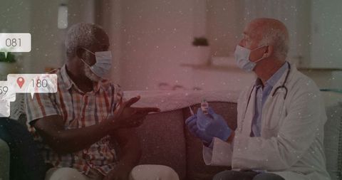 Doctor explaining home vaccination to masked senior patient on sofa, gloved holding vial