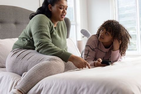 Multicultural women reviewing paperwork on bed, discussing finances and using smartphone