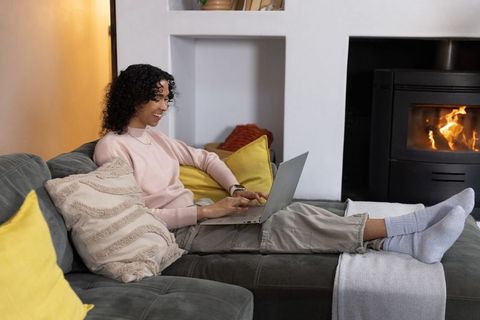 Non-binary person working from home on laptop reclining on cozy gray sofa beside fireplace