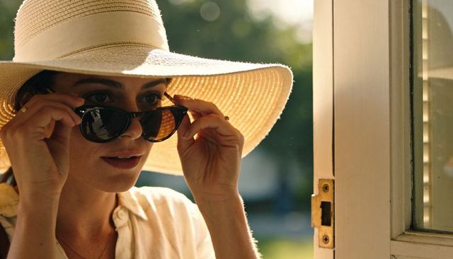 Adjusting sunglasses wearing wide-brim straw hat at sunlit doorway, chic summer style