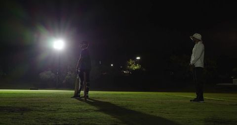 Night cricket batter standing at stumps under floodlight casting dramatic long shadow
