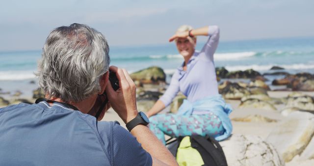 Older couple enjoying sunny day taking photos on the beach. Woman posing with hand shielding eyes from sunlight. Ideal for themes about travel, leisure, outdoor activities, and senior lifestyles.