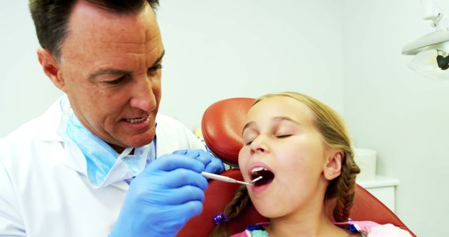 Dentist examining a young girl's teeth during a dental appointment. The dentist is using dental tools while the young girl sits in the dental chair with her mouth open. Ideal for use in articles or advertisements about dental care, pediatric dentistry, children's healthcare, and dental check-ups.