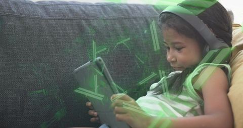 Young girl reclining on sofa using tablet with headphones, green digital overlay learning