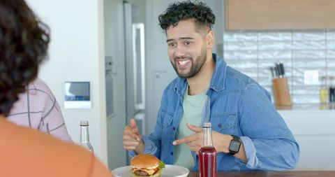 Bearded hispanic man pointing at cheeseburger while chatting with friends at kitchen counter