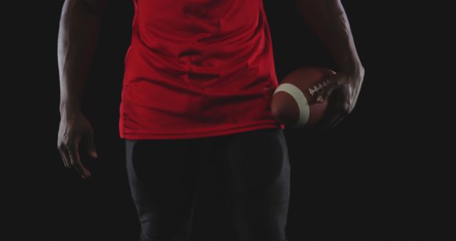 Athletic man holding a football, dressed in red shirt and black pants, under a dark background. Perfect for sports advertising, fitness promotions, football game posters, and athletic training programs.