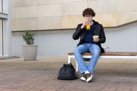 Young man sitting on bench drinking orange juice while eating sandwich on campus plaza