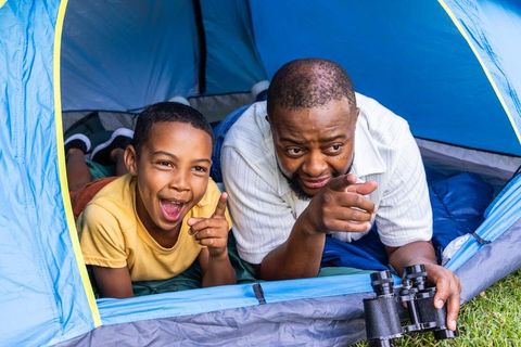 African american father and teen son camping and bonding in blue tent pointing at wildlife