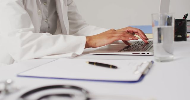 Doctor typing on a laptop at an office desk, with medical tools like a stethoscope and clipboard present. Could be used for themes related to healthcare, medical research, telemedicine, digital health records, or professional work environments in the medical field.