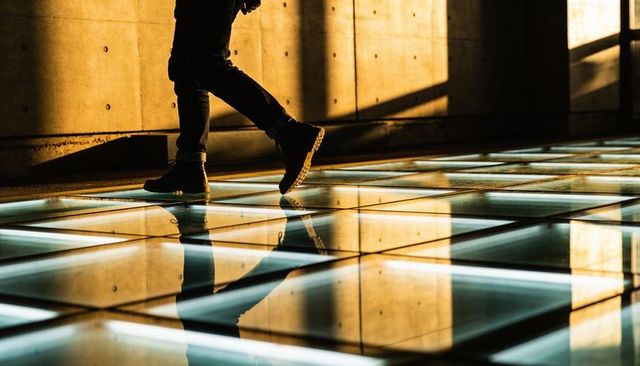 Walking man crossing illuminated glass floor with bold shadow and concrete wall reflection