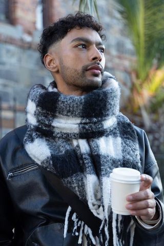 Urban young man in leather jacket wearing plaid scarf holding coffee cup, moody portrait