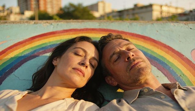 Middle-aged couple resting on rooftop wall with rainbow mural at golden hour