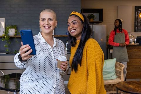Smiling diverse women taking selfie in cozy cafe with coffee cup and vibrant knitwear