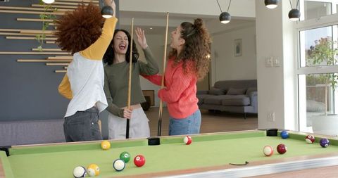 Three women celebrating while holding cue sticks around pool table in modern living room