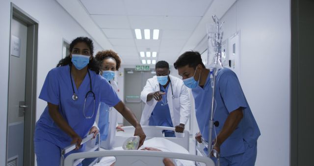 Doctors and nurses in medical uniforms are urgently pushing a patient's bed down a hospital hallway. The healthcare workers, wearing masks, appear focused and determined, indicating an emergency situation. This can be used to illustrate themes of medical emergency, teamwork in healthcare, and quick response scenarios in hospital settings.
