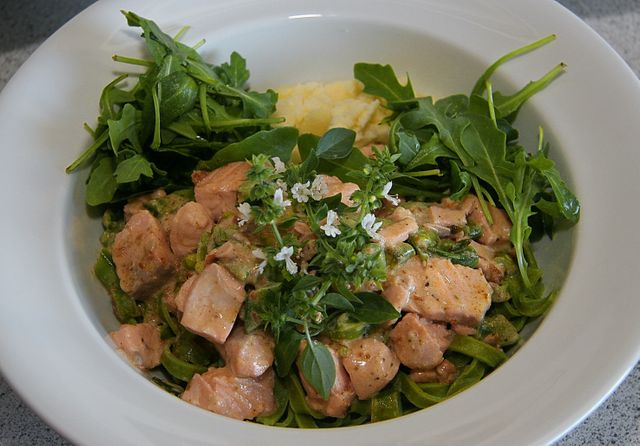 Salmon pasta topped with green herbs, garnished with fresh arugula and served in a white bowl on a neutral surface. Ideal for use in websites and blogs about healthy eating, gourmet recipes, food photography, and meal preparation guides.