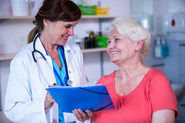 Senior woman consulting with a doctor in a hospital setting. The doctor is holding a clipboard and both are smiling, indicating a positive interaction. This image can be used for healthcare websites, medical brochures, articles about elderly care, and promotional materials for hospitals and clinics.