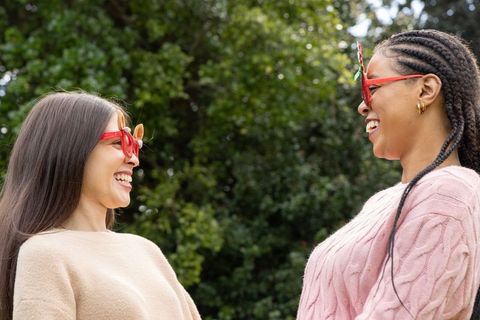 Diverse friends laughing outdoors wearing red novelty glasses, knit sweaters, braids
